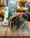 Blue teddy bear with a bouquet of flowers and a baby blanket on a white background