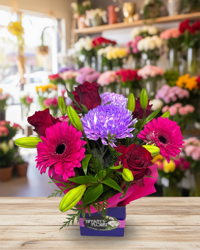 Bouquet of colorful flowers including pink gerberas, red roses, and purple asters in a box with 'The Faintest Flower' branding.