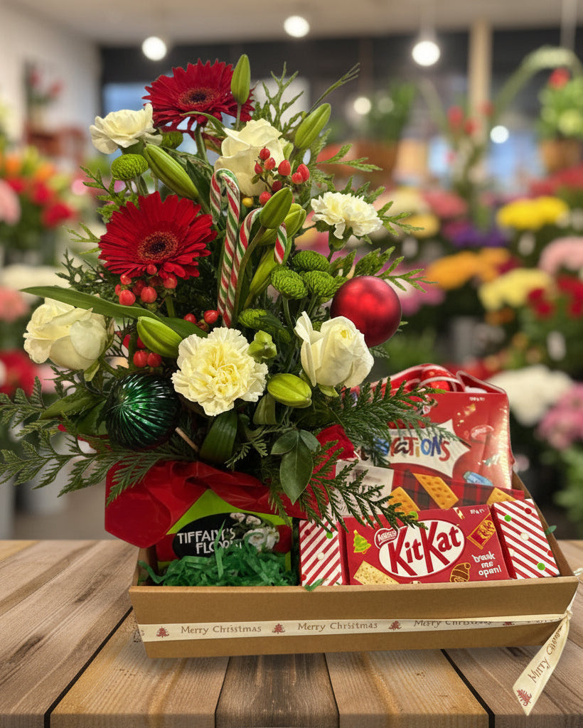 A festive Christmas gift basket filled with a red and white floral arrangement, candy canes, greenery, ornaments, and assorted holiday treats including KitKat, Tifani's Pecan Butter, and Reese's, presented in a brown box with a ribbon.