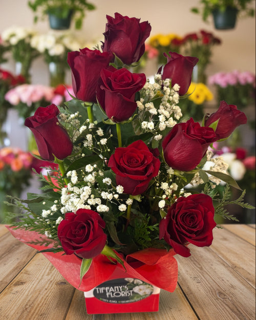 Bouquet of red roses with greenery and white flowers in a box labeled Tiffany's Florist on a white background.